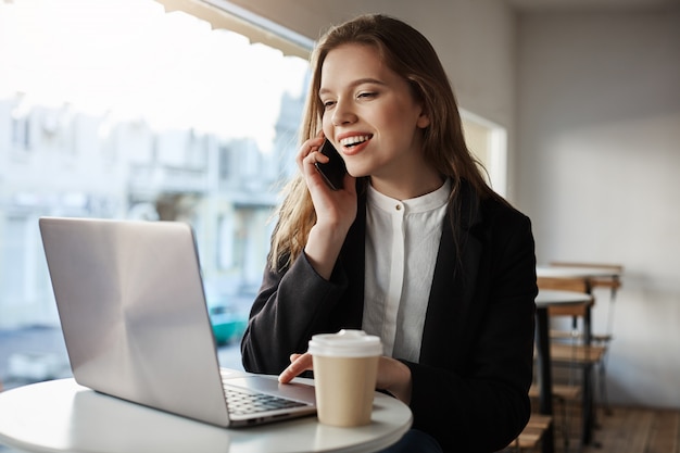 donna caucasica seduto nella caffetteria, bere caffè, parlare su smartphone, guardando lo schermo del laptop con ampio sorriso