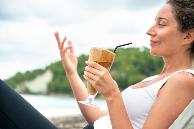 Donna caucasica sorridente che tiene la bevanda del caffè su una spiaggia con schiuma e cannuccia con le colline