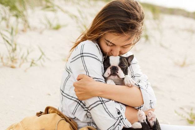 Donna che abbraccia il suo bulldog sulla spiaggia nella luce del tramonto, vacanze estive. Ragazza alla moda con cane divertente che riposa, abbraccia e si diverte, momenti carini.