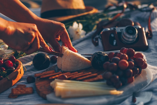 donna che affetta formaggio sul tagliere di legno con formaggio e frutta su di esso e fotocamera, cappello e fiori in spiaggia.