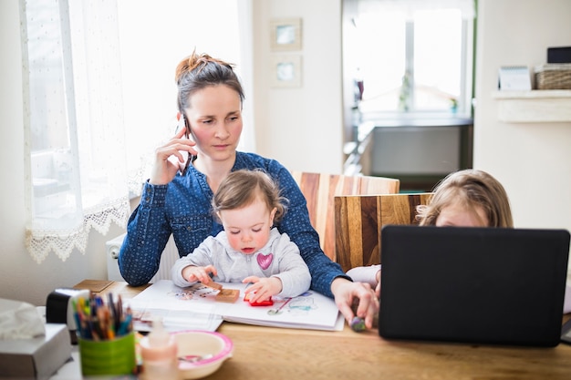 Donna che comunica sullo smartphone mentre i suoi bambini che giocano sopra lo scrittorio