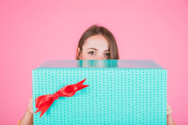 Donna che dà una occhiata dal grande contenitore di regalo con l&#39;arco rosso contro fondo rosa