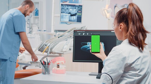 Donna che guarda lo schermo verde sul telefono cellulare per cure stomatologiche presso l'ufficio di odontoiatria. Ortodontista che tiene smartphone con chiave cromatica e modello mockup in esposizione per la cura dei denti