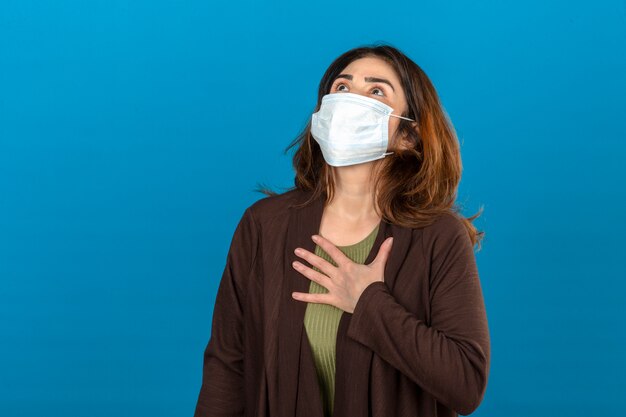 Donna che indossa cardigan marrone in maschera protettiva medica toccando sul petto per controllare il polmone mentre si respira sopra la parete blu isolata