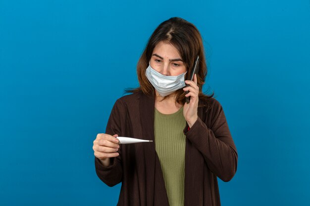 Donna che indossa cardigan marrone in smartphone protettivo medico della tenuta della maschera che esamina termometro digitale a disposizione che chiama a qualcuno che sembra nervoso sopra la parete blu isolata