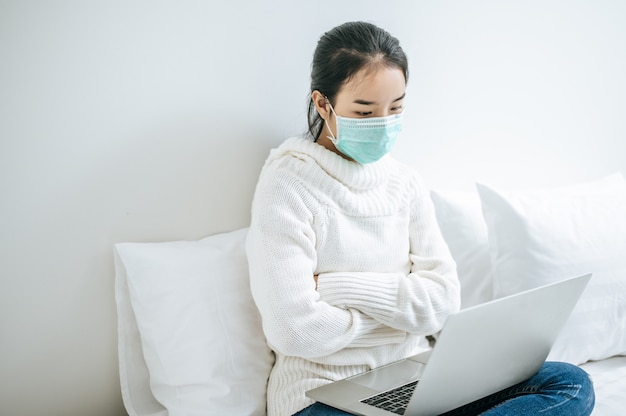 Donna che indossa una maschera, seduto sul letto e giocare a laptop.