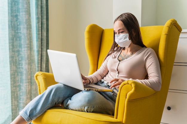 Donna che lavora al computer portatile dalla poltrona durante la pandemia mentre indossa la maschera medica