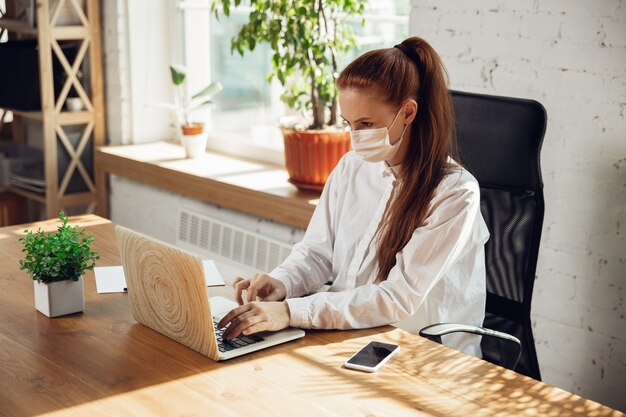 Donna che lavora da sola in ufficio durante la quarantena di coronavirus o covid con maschera facciale