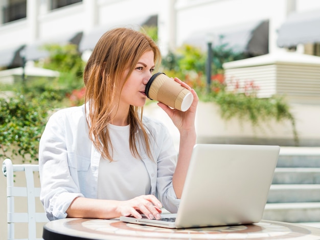 Donna che mangia caffè all'aperto mentre lavorando al computer portatile