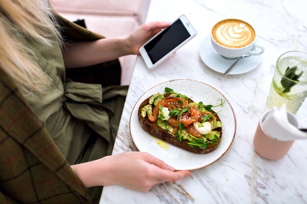 Donna che mangia il suo gustoso brunch sul caffè hipster, vista dall'alto del tavolo di marmo, toast di salmone e avocado, caffè e dolci gustosi cheesecake, gustando la sua colazione.