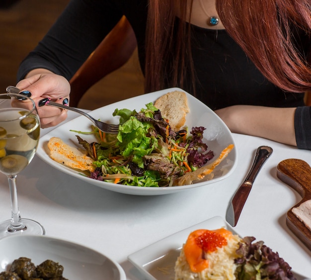 Donna che mangia insalata verde con patatine fritte e un bicchiere di prosecco.