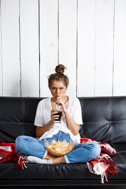 donna che mangia patatine, bere soda, guardare la tv, seduto al divano.