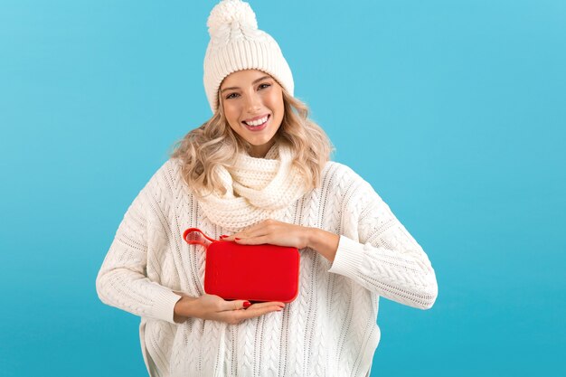 Donna che mantiene l'altoparlante wireless ascoltando musica felice indossando un maglione bianco e berretto lavorato a maglia in posa isolato su blue