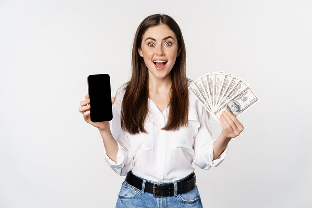 Donna che mostra lo schermo del telefono cellulare e contanti, denaro, concetto di microcredito e prestiti bancari, in piedi su sfondo bianco.