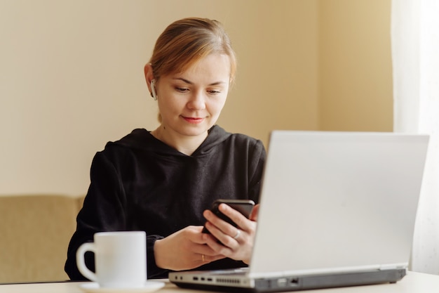 Donna che per mezzo del computer portatile e del telefono cellulare per il suo lavoro a distanza a casa