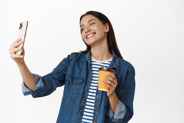 donna che prende selfie con una tazza di caffè da asporto sorridente felice, giornata rilassante, in piedi su bianco