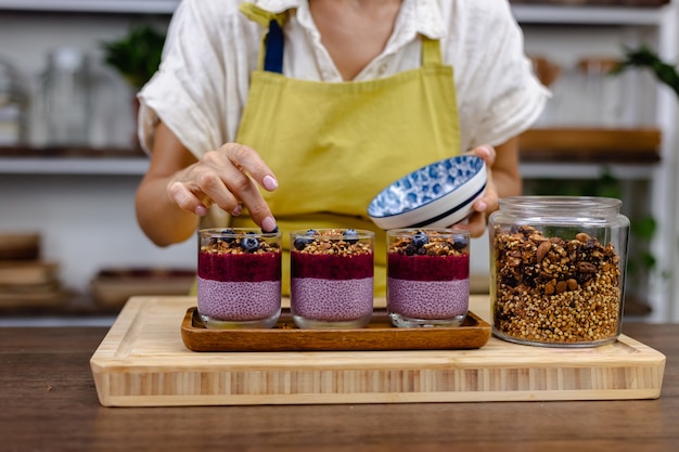 Donna che prepara deliziosi budini di chia del deserto con fragole e mirtilli, latte di mandorle con polvere di rosa di frutta del drago e muesli in cucina a casa.