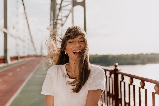 Donna che ride eccitata con un sorriso meraviglioso che indossa una maglietta bianca se gli occhi chiusi e sorride e si gode la mattina d'estate camminando sul ponte in città