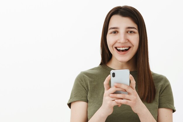 Donna che ride sulla foto divertente di te scattata tramite smartphone tenendo il telefono cellulare in mano sorridendo ampiamente divertito e spensierato divertendosi come posa contro il muro bianco con dispositivo