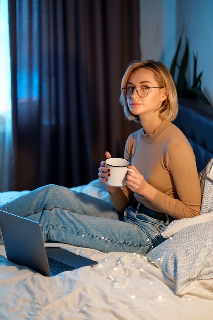 Donna che si distende e che beve tazza di caffè o tè caldo utilizzando il computer portatile in camera da letto.