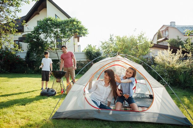 Donna che si siede con sua figlia in tenda durante il marito e il figlio che cucinano sulla griglia del barbecue