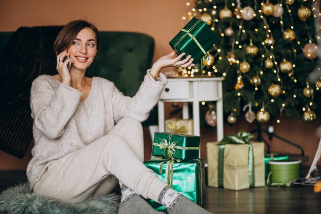 Donna che si siede dall'albero di Natale e che compera sul telefono