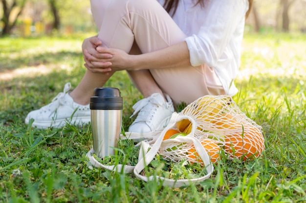 Donna che si siede sull'erba con la borsa e il thermos riutilizzabili