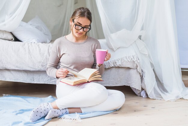 Donna che si siede vicino al letto che beve caffè durante la lettura del libro