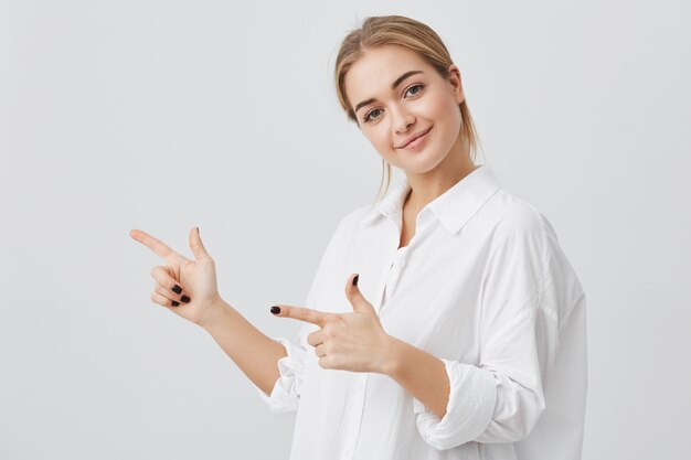 Donna che sorride allegramente e puntando il dito indice, mostrando copia spazio per il testo o il contenuto pubblicitario. Colpo dello studio di giovane ragazza bionda attraente isolata su fondo grigio