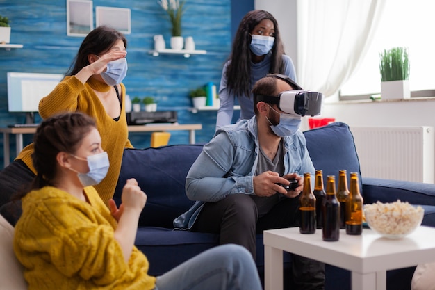 Donna che sperimenta la realtà virtuale giocando ai videogiochi con l'auricolare vr che indossa una maschera facciale mentre gli amici si rallegrano mantenendo le distanze sociali indossando una maschera facciale per prevenire l'infezione da virus, birra