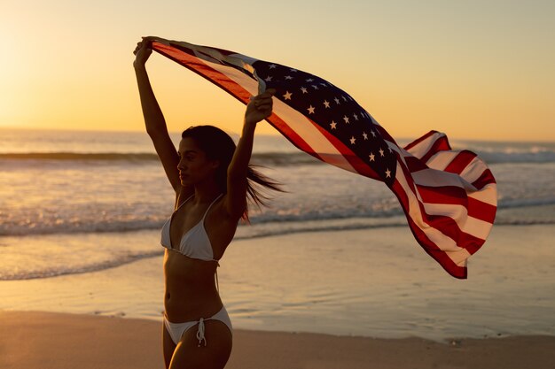 Donna che sta con la bandiera americana d'ondeggiamento sulla spiaggia