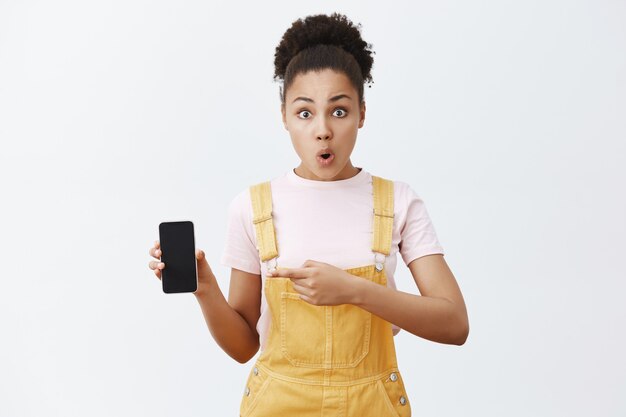 donna che tiene smartphone e punta allo schermo con il dito indice, piegando le labbra dall'interesse e dalla sorpresa, in piedi scioccato e impressionato