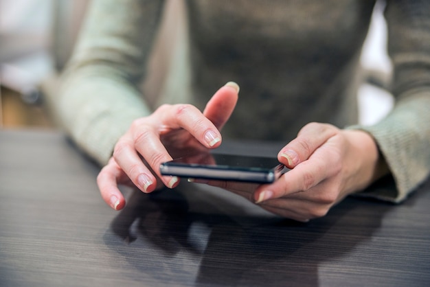 Donna che tiene uno smartphone sul tavolo di legno. Primo piano della ragazza, seduta al tavolo di legno, in una mano è smartphone. Imprenditrice navigare su internet su smartphone.