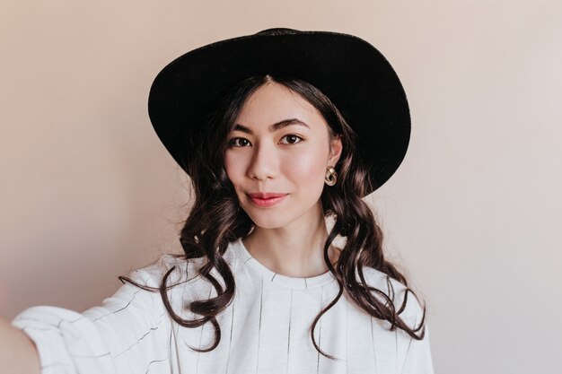 Donna cinese riccia che cattura selfie. Vista frontale del modello asiatico in cappello in posa su sfondo beige.