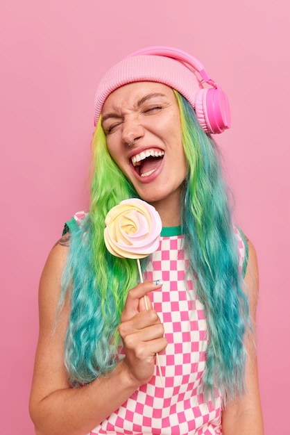 donna con acconciatura colorata alla moda ascolta musica tramite cuffie wireless si diverte tiene caramelle rotonde su bastone indossa cappello e abito a scacchi isolato su rosa
