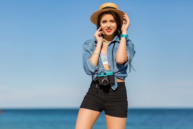 donna con corpo abbronzato, labbra rosse piene el gambe lunghe in posa sulla spiaggia soleggiata tropicale. Indossare top corto, pantaloncini e cappello di paglia.