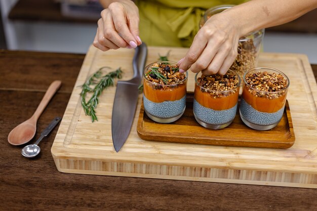 Donna con dolci sani e colorati budini di chia a base di latte di mandorle, estratto di spirulina blu, semi di chia, marmellata di mango pappaya e muesli fatto in casa. Sul tavolo di legno in cucina a casa.