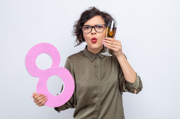 Donna con i capelli corti che tiene il numero otto realizzato in cartone e bicchiere di champagne guardando la telecamera sorpresa celebrando la giornata internazionale della donna l'8 marzo in piedi su sfondo bianco