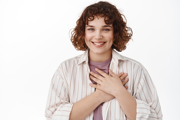 donna con i capelli corti e ricci, tiene le mani sul cuore e sorride felice alla telecamera, ringraziandoti, apprezza il bel gesto, essendo grata, in piedi sul bianco.