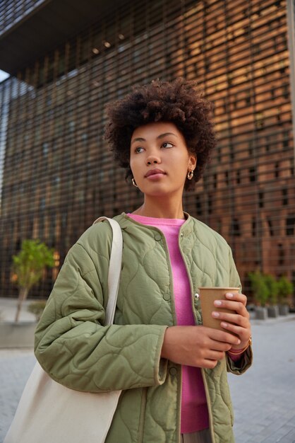 donna con i capelli ricci beve caffè da un bicchiere di carta porta una borsa vestita in abiti casual si trova all'aperto cammina e si gode una bella giornata durante la ricreazione