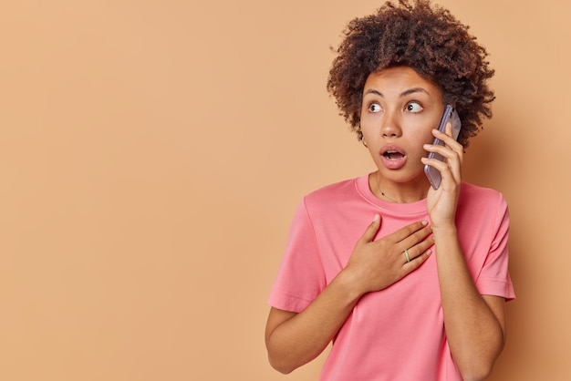 donna con i capelli ricci parla al telefono ascolta informazioni sente notizie incredibili con espressione sorpresa tiene la mano sul petto concentrata lontano pose contro beige