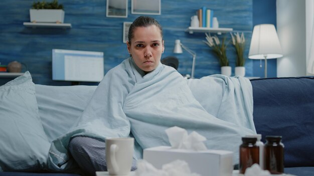 Donna con infezione da virus seduta in coperta guardando la fotocamera. Persona malata che sente freddo e trema a causa dell'influenza stagionale a casa. Adulto con malattia e trattamento contro la malattia