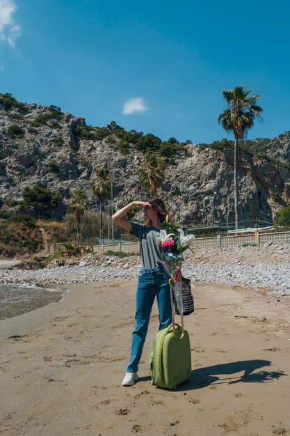 Donna con la borsa dei bagagli e mazzo del fiore che protegge il suo occhio che sta sulla spiaggia