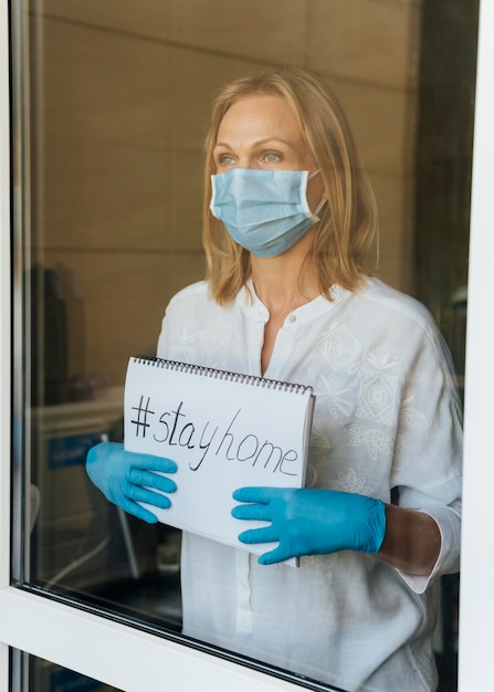 Donna con la mascherina medica che tiene il taccuino domestico di soggiorno alla finestra