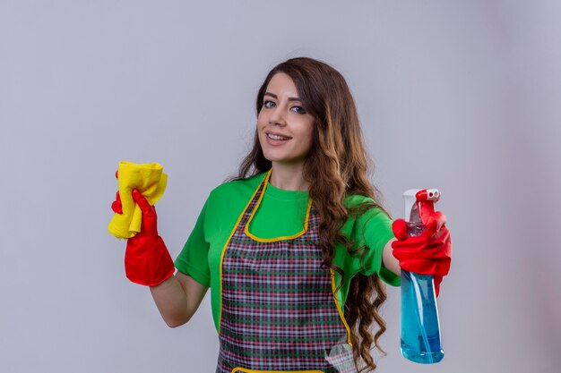 donna con lunghi capelli ondulati che indossa grembiule e guanti di gomma che tiene tappeto e spray per la pulizia guardando positivo sorridente fiducioso in piedi