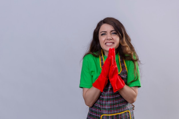 donna con lunghi capelli ondulati che indossa grembiule e guanti di gomma elemosinando e pregando con le mani insieme con l'espressione di speranza sul viso in piedi