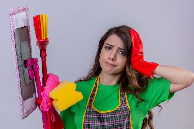 donna con lunghi capelli ondulati che indossa grembiule e guanti di gomma in piedi con strumenti di pulizia e stracci toccando la testa guardando triste stanco di pulizia