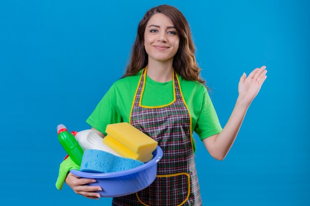 donna con lunghi capelli ondulati che indossa grembiule e guanti di gomma tenendo il bacino con il pedaggio di pulizia sorridente agitando con la mano in piedi sul blu