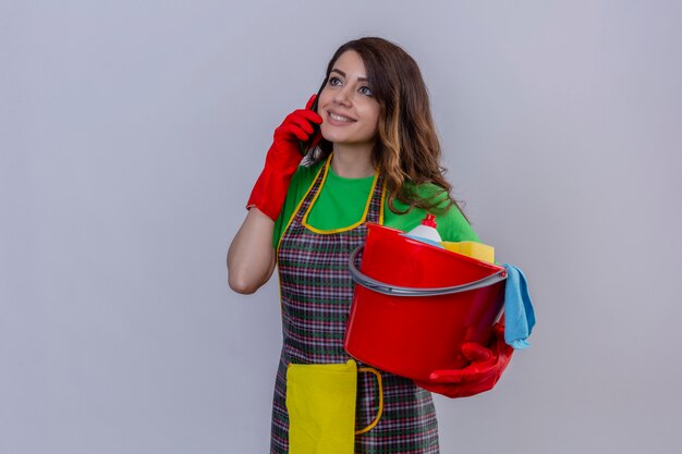 donna con lunghi capelli ondulati in grembiule e guanti che tiene secchio con strumenti di pulizia mentre parla al telefono cellulare sorridente in piedi