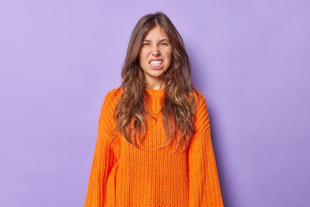 donna con lunghi capelli scuri stringe i denti con irritazione strizza gli occhi faccia infastidita da qualcosa indossa un maglione lavorato a maglia arancione casual isolato su viola